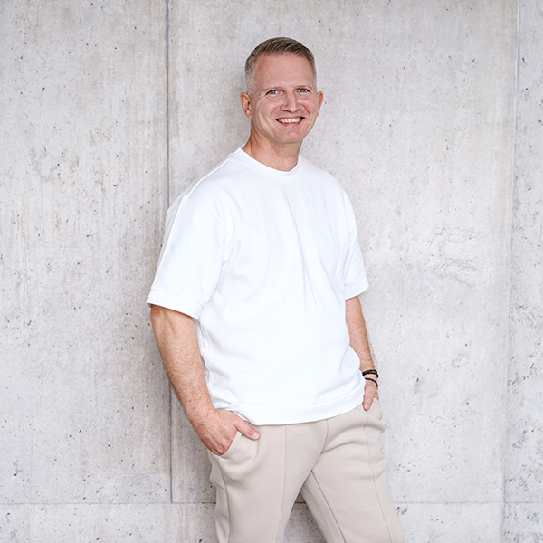 Frank Gohl lächelnd in weißem T-Shirt und beiger Hose steht mit den Händen in den Taschen vor einer hellgrauen Betonwand.
