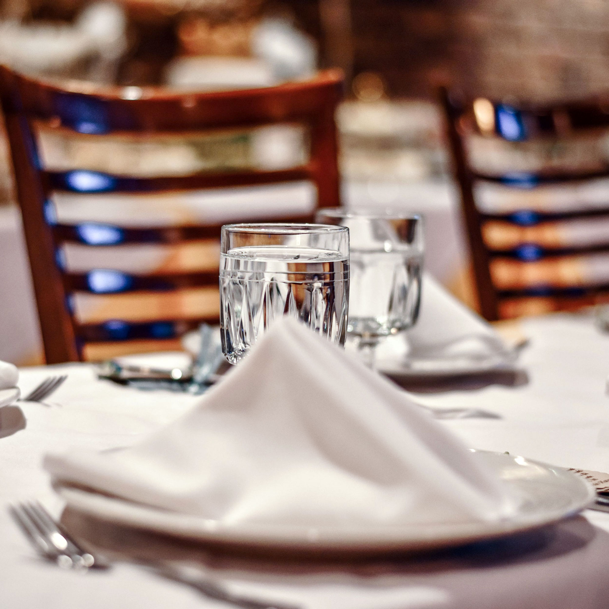 Glas mit Wasser auf gedecktem Tisch mit weißer Serviette und Holzstühlen im Hintergrund