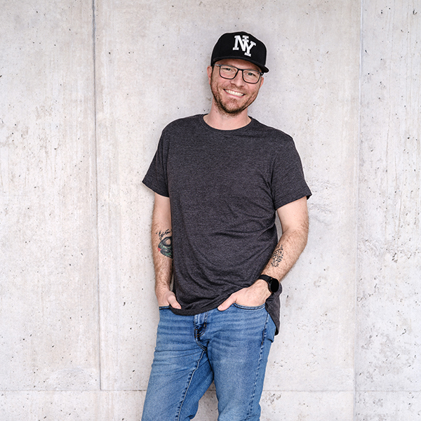 Herr Dennis Hinel mit Brille, schwarzer NY-Kappe, grauem T-Shirt und blauen Jeans steht lächelnd mit den Händen in den Taschen vor einer hellen Betonwand.
