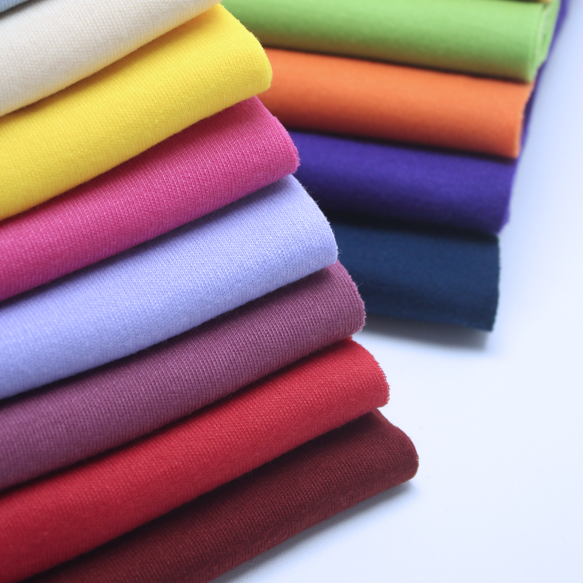 Gestapelte Stoffstücke in Gelb, Pink, Hellblau, Violett, Rot, Dunkelrot, Grün, Orange und Dunkelblau auf hellem Untergrund.