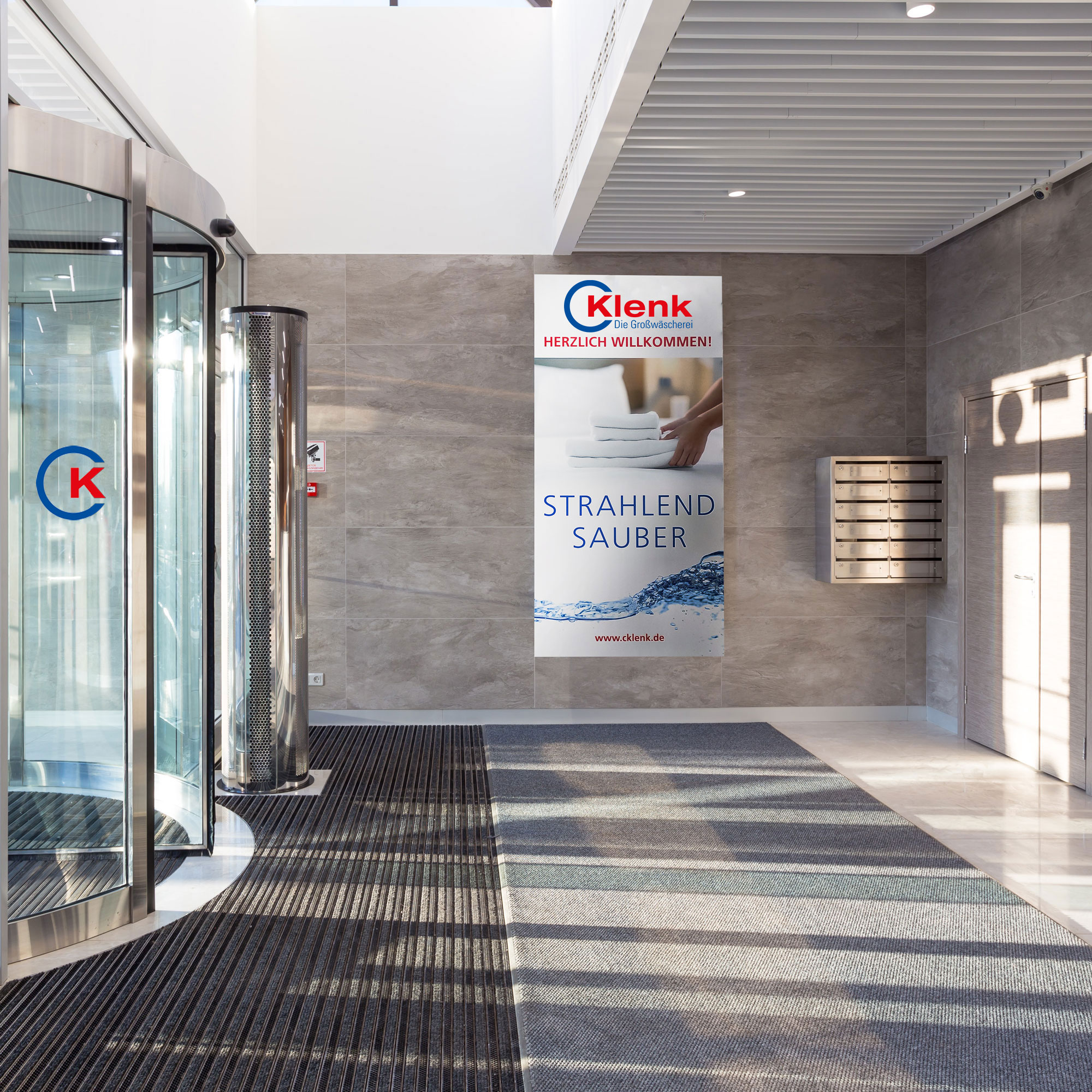 Eingangsbereich mit Drehkreuz und Schmutzfangmatten, grauem Bodenbelag und Werbeplakat der Firma Klenk mit Text 'Herzlich willkommen! Strahlend sauber'.