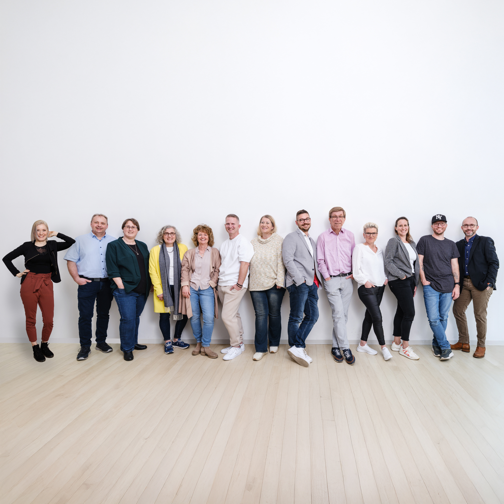 Das Team der Verwaltung steht in einer Reihe vor einer weißen Wand, trägt legere Kleidung in verschiedenen Farben und posiert entspannt.