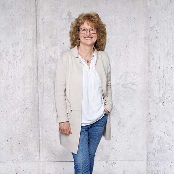 Frau Silvia Storz in Jeans und weißem Hemd steht vor einer strukturierten Betonwand.