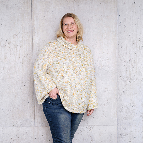 Frau Nadine Mattausch mit blonden Haaren, die einen hellen Strickpullover und Jeans trägt, steht lächelnd vor einer hellgrauen Betonwand.