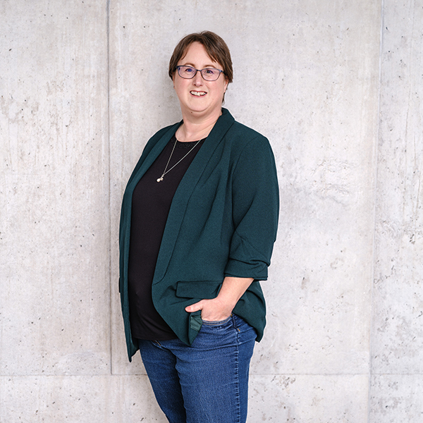 Frau Diana Bauer in grüner Jacke, schwarzem Oberteil und blauer Jeans steht vor einer Betonwand, eine Hand in der Hosentasche.