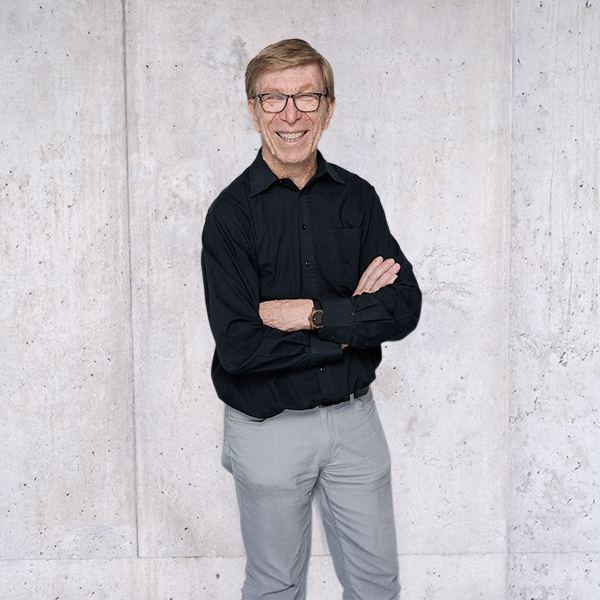 Herr Thomas Traub mit Brille und schwarzem Hemd steht vor einer schlichten Wand und blickt direkt in die Kamera.