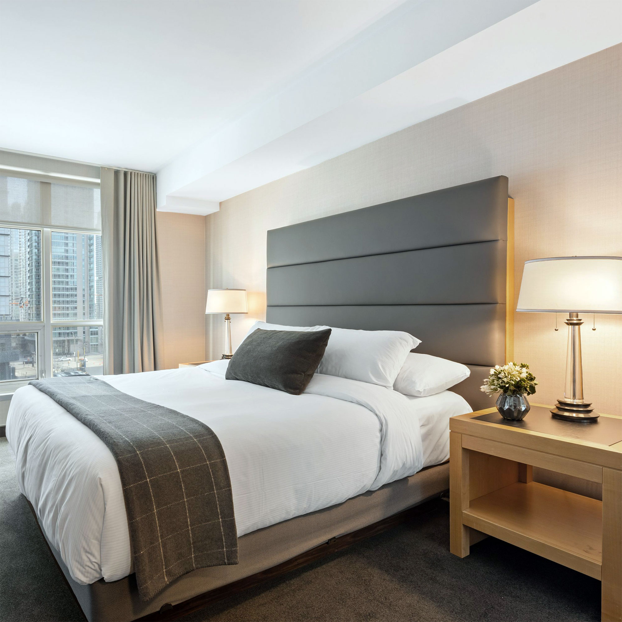 Modern eingerichtetes Hotelzimmer mit großem Bett, grauem Kopfteil, weißen Bettwäsche und grauer Decke, zwei Nachttischen mit Lampen und Fenster mit Blick auf Hochhäuser.