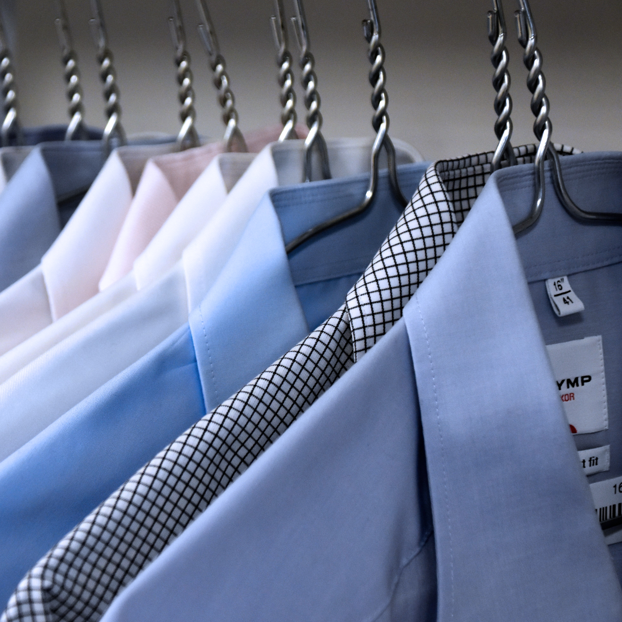 Hemden verschiedener Farben und Muster hängen auf Metallbügeln, darunter hellblaue, weiße und kariert gemusterte Hemden.