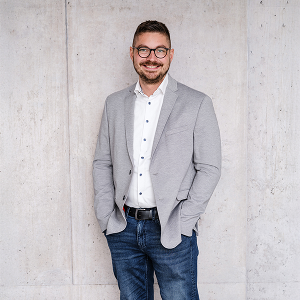 Herr Pascal Henke mit Brille, der einen hellgrauen Blazer, ein weißes Hemd und blaue Jeans trägt, steht vor einer hellen Betonwand.