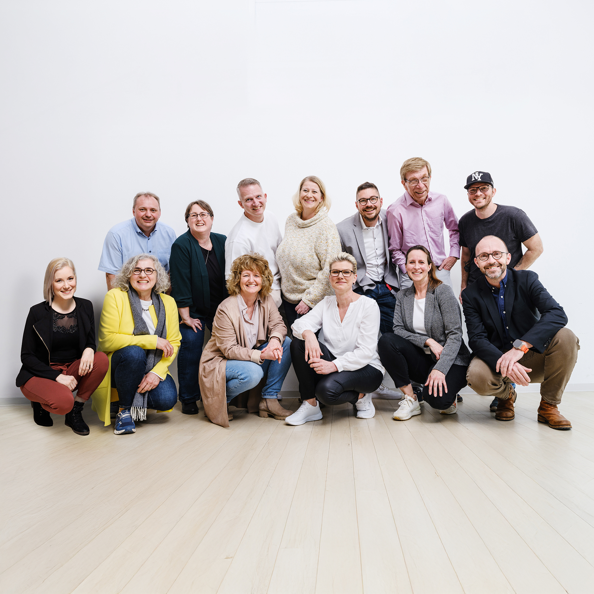 Das Team der Verwaltung in legerer Kleidung, einige sitzend, andere stehend, vor weißer Wand auf hellem Holzboden.