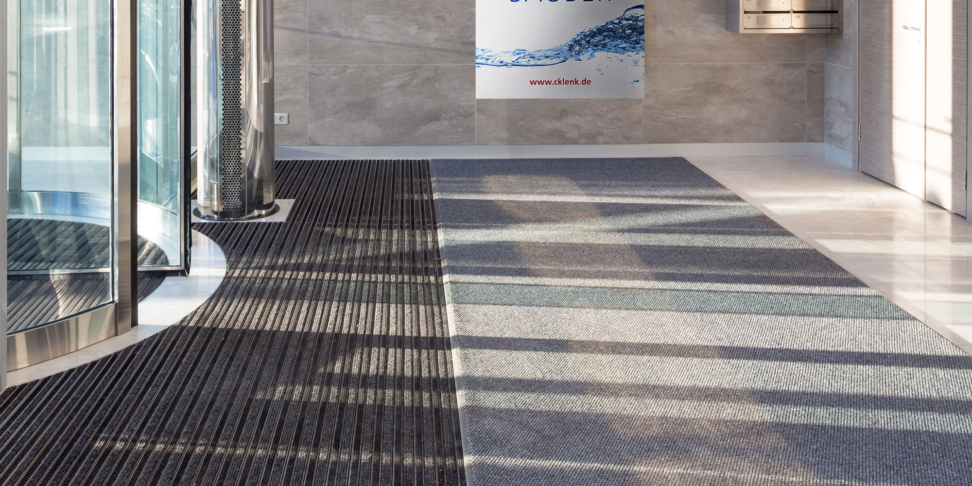 Eingangsbereich mit Drehkreuz und Schmutzfangmatten, grauem Bodenbelag und Werbeplakat der Firma Klenk mit Text 'Herzlich willkommen! Strahlend sauber'.